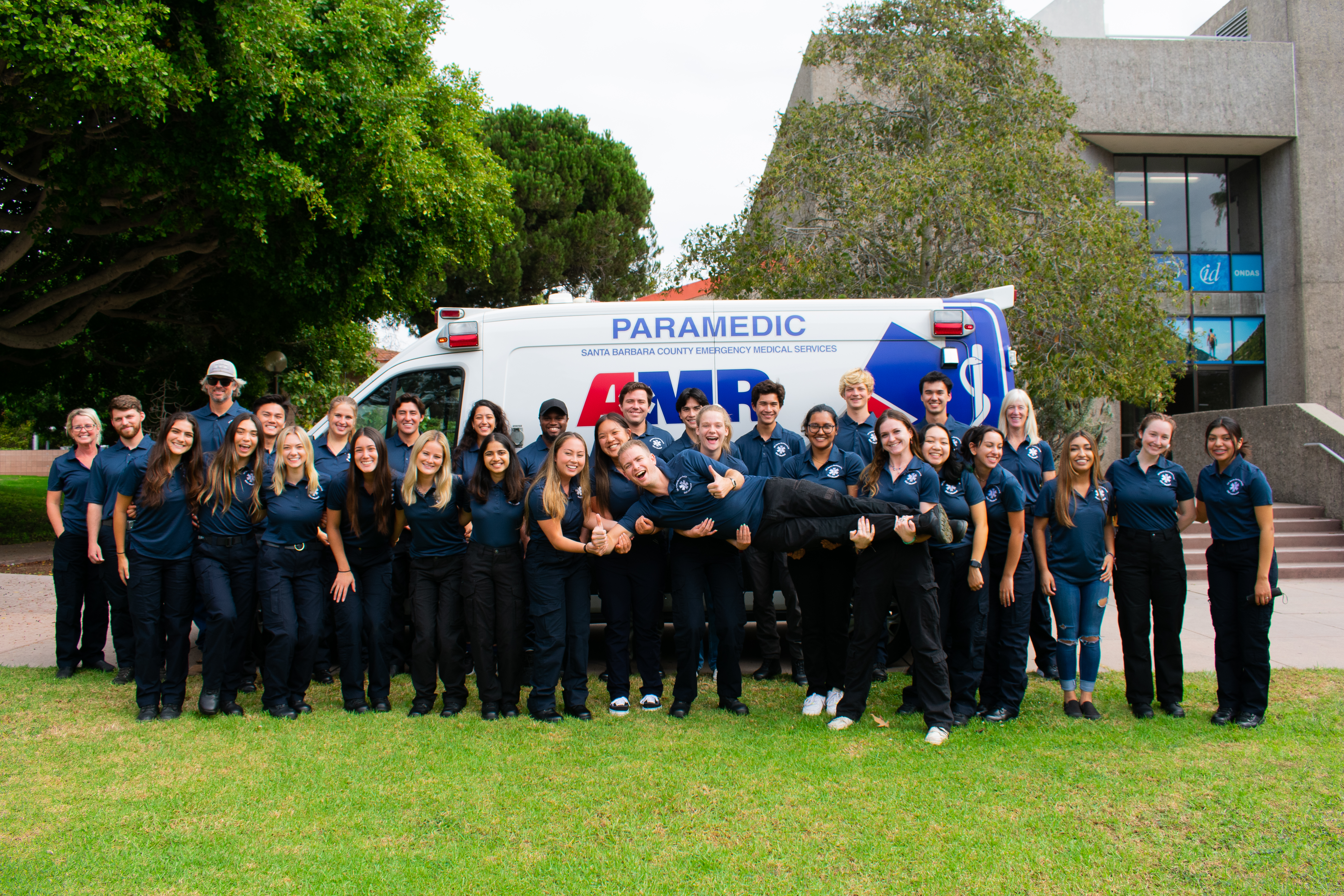 EMT Class standing in their navy blue EMT shirts in front of an AMR ambulance, holding up their lead instructor on their arms